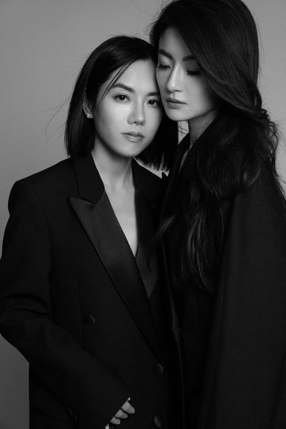 Black and white portrait of two women with dark hair, standing close and facing the camera, wearing formal black jackets.