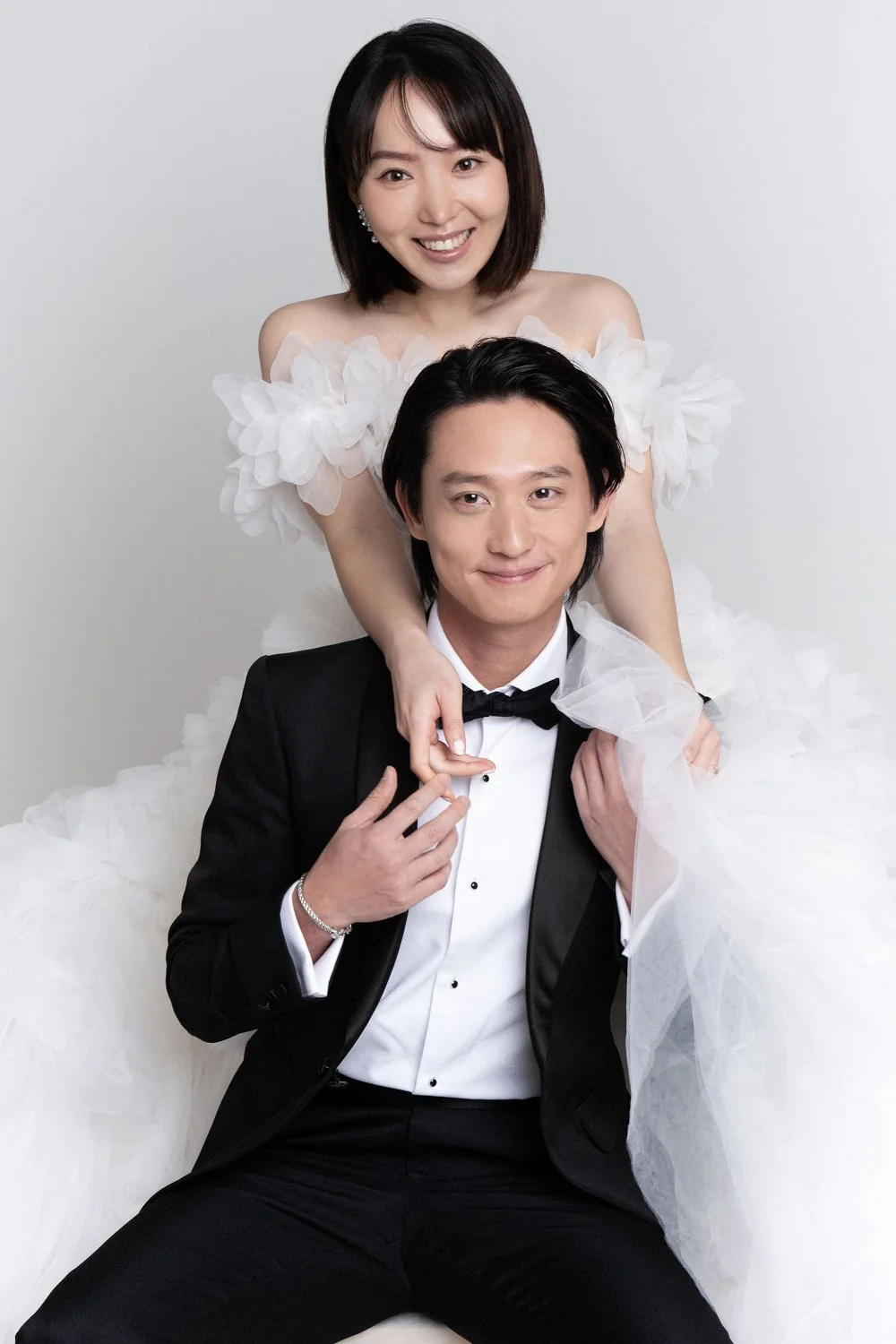 A woman in a white off-shoulder dress with feather details and a man in a tuxedo with a bow tie, both smiling, posing for a formal portrait against a plain background.