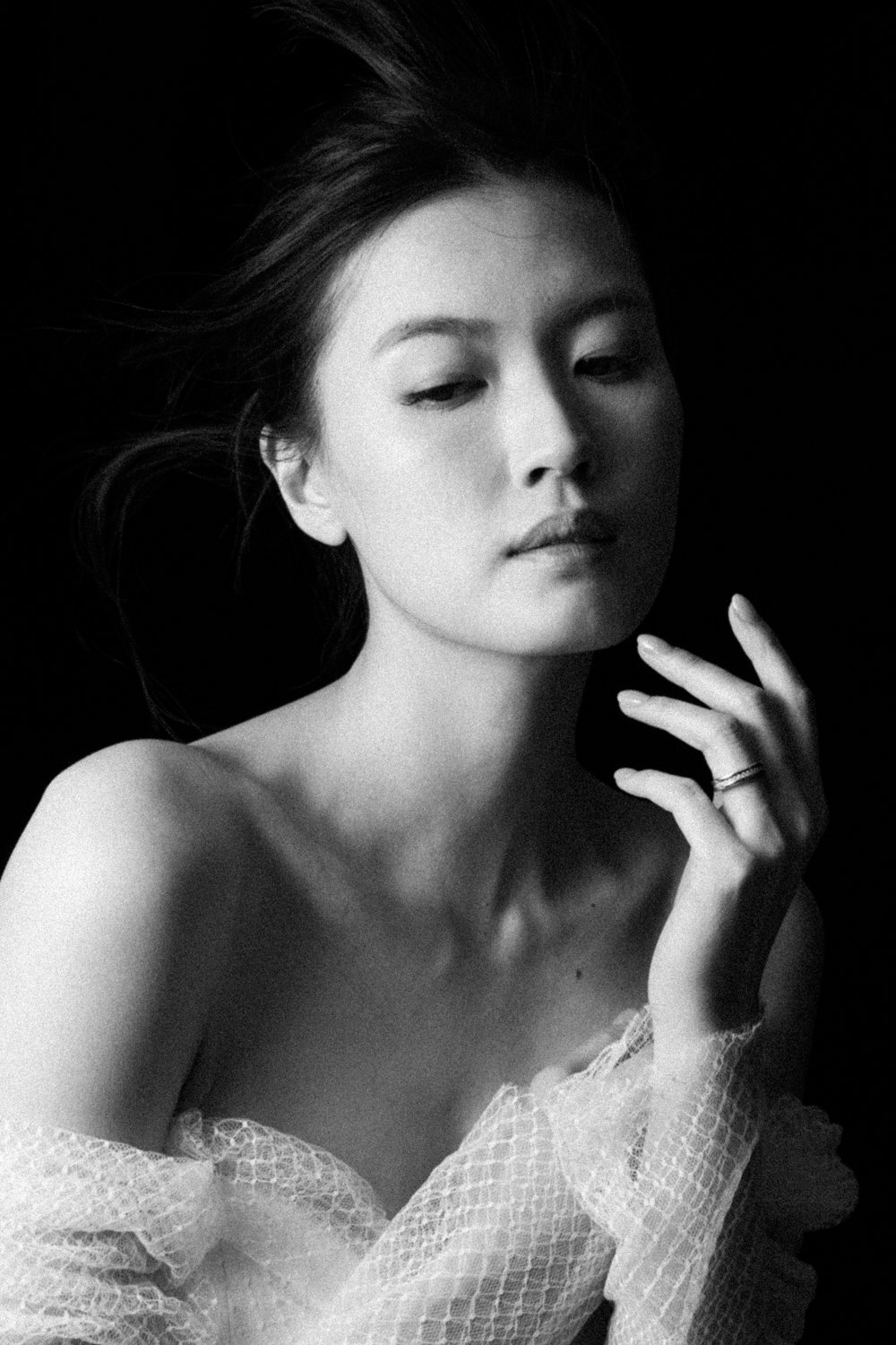 A pregnant woman with shoulder-length brown hair, wearing a satin beige dress, standing against a dark background, holding her belly with one hand and looking at the camera.