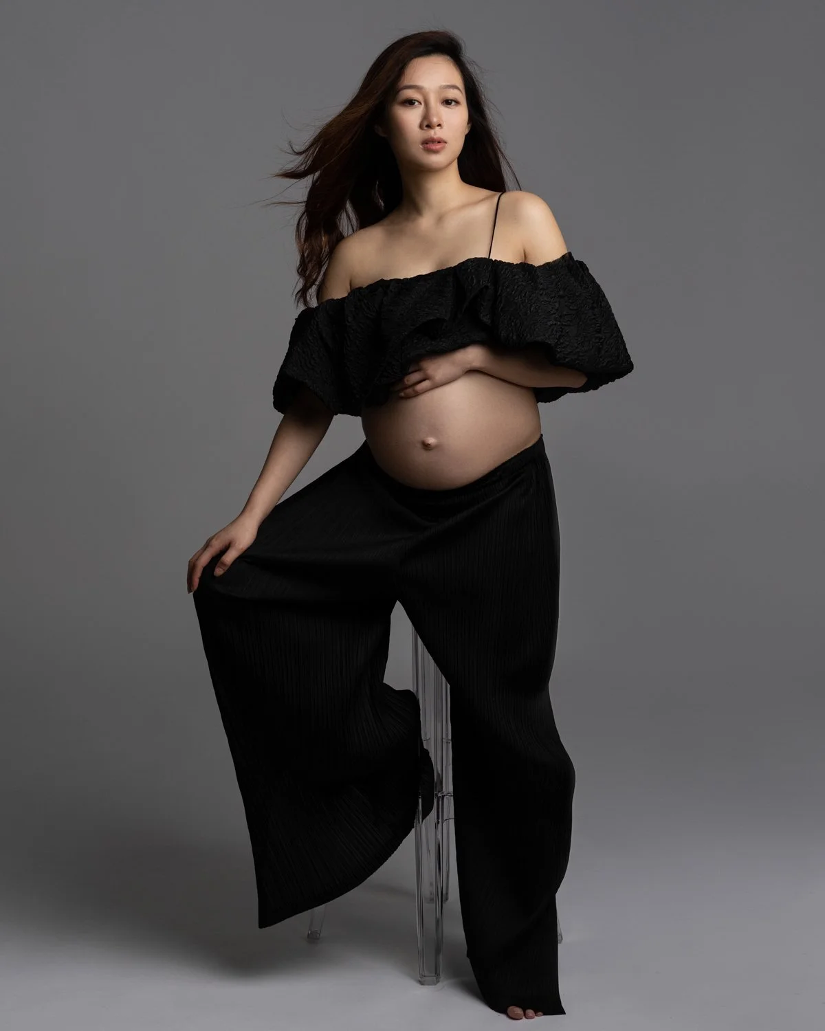 A pregnant woman with long dark hair, wearing an off-shoulder black top and black wide-leg pants, posing against a gray background.