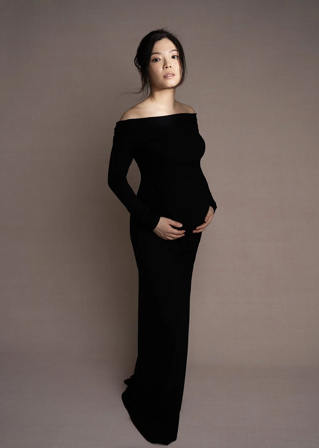 A pregnant woman in a black off-the-shoulder dress posing against a plain brown background.