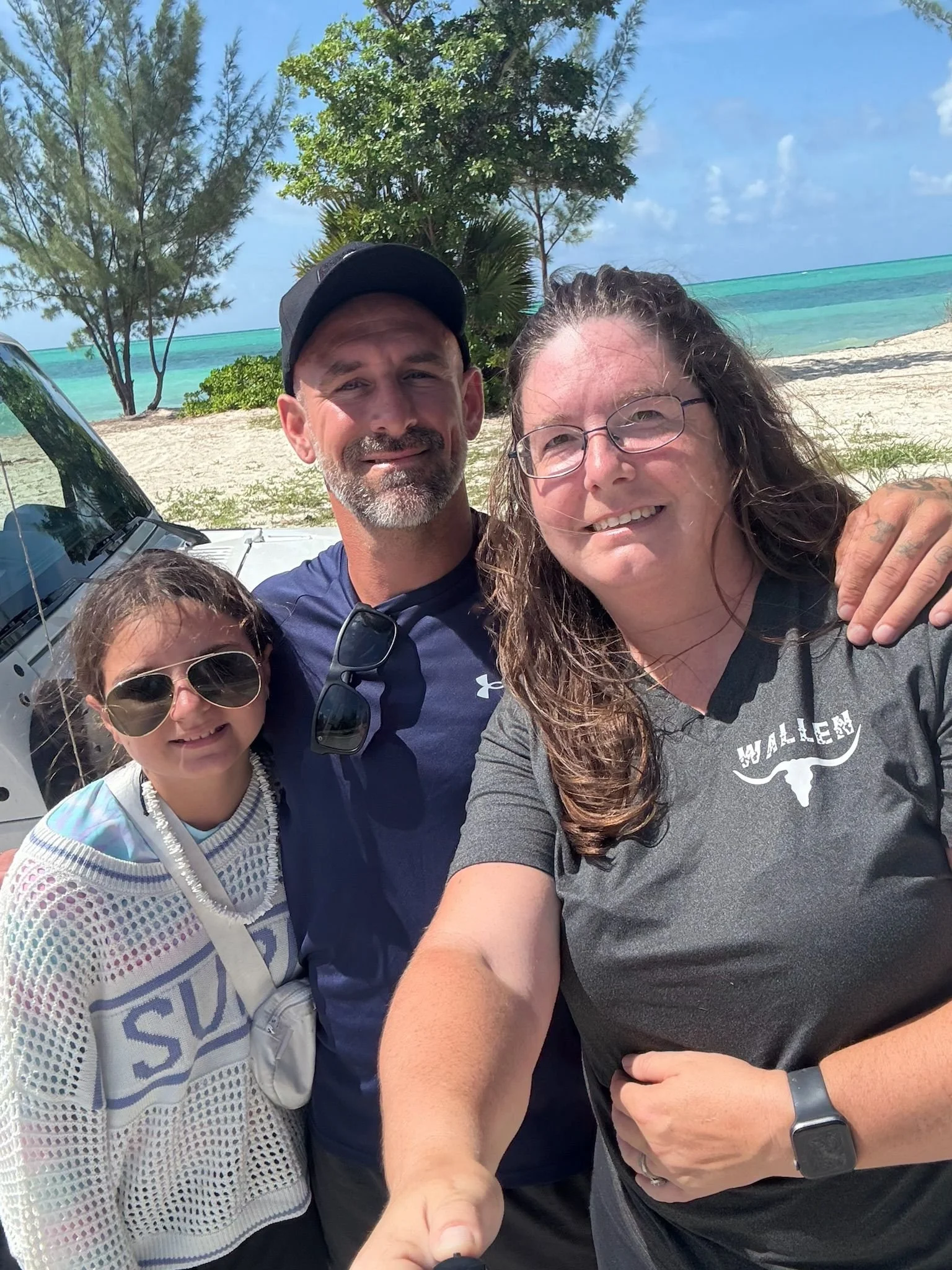 A family of three taking a selfie on a beach with turquoise water and trees in the background. The man is in the middle, the woman on the right, and the girl on the left wearing sunglasses.