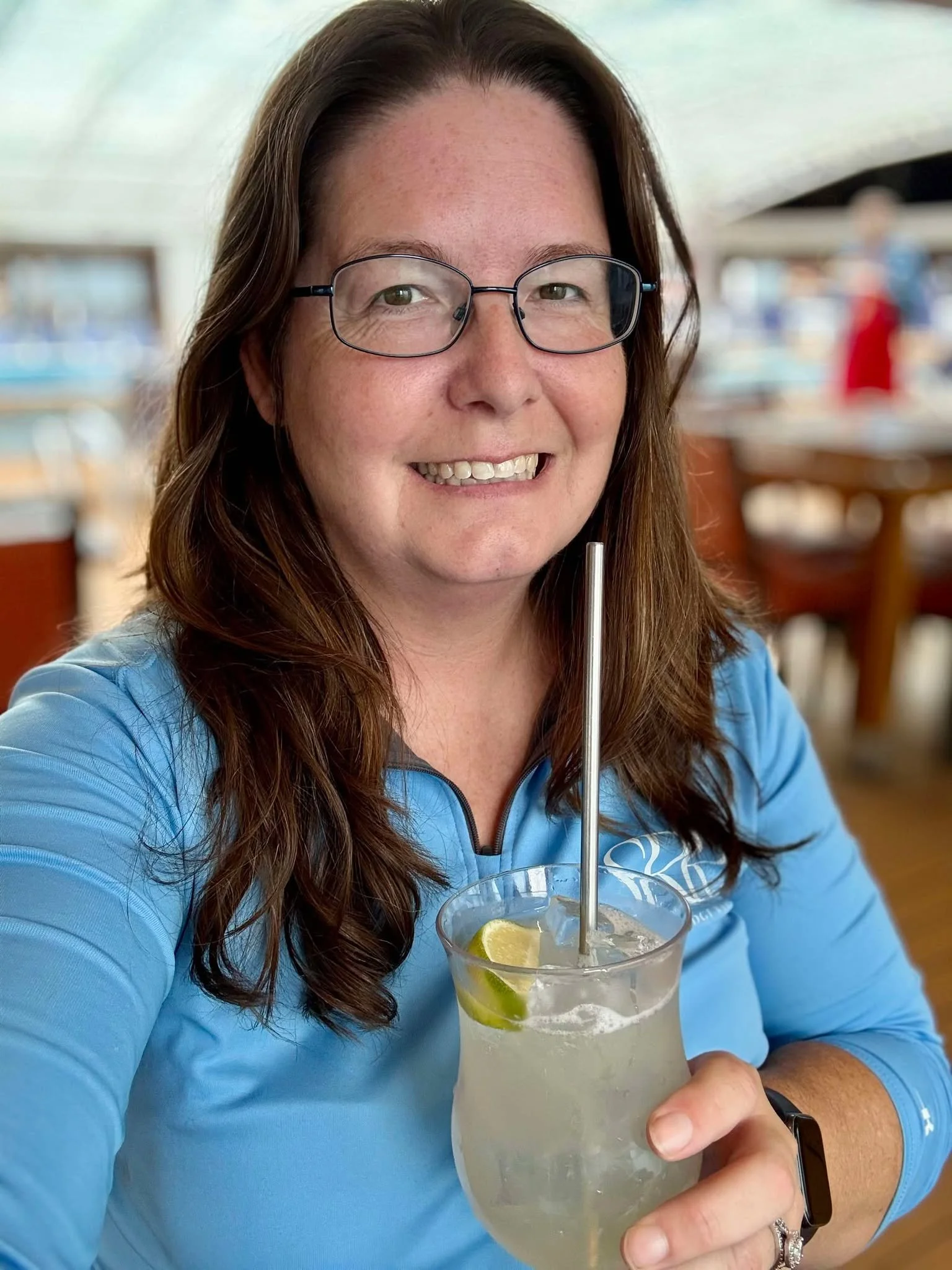 A woman with long brown hair, glasses, and a blue jacket smiling and holding a glass of lemonade with lemon slices and a straw.