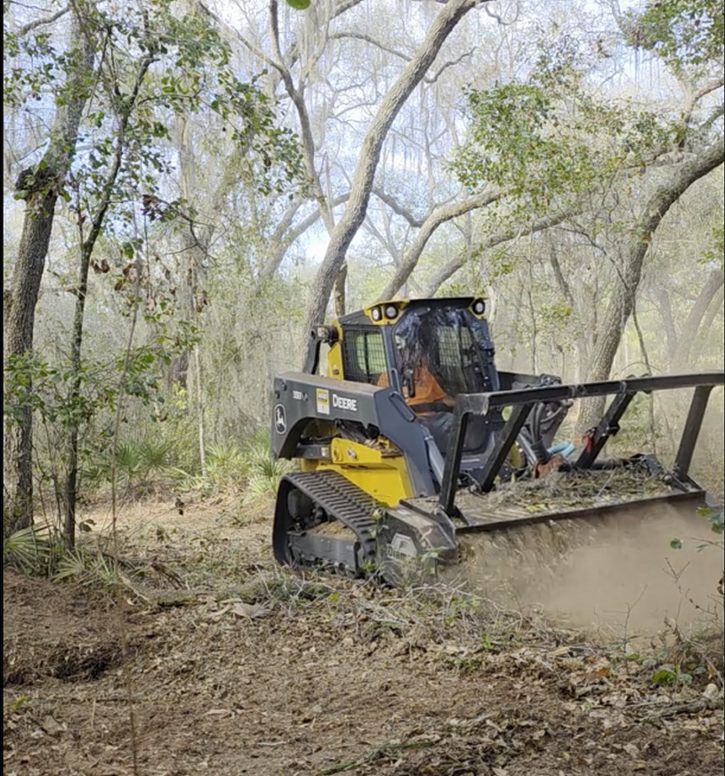 A John Deere skid steer loader clearing brush and debris in a wooded area.