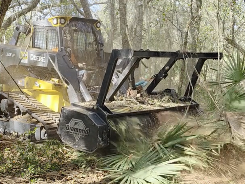 A John Deere compact track loader operating a land clearing attachment in a wooded area, with trees and branches around.