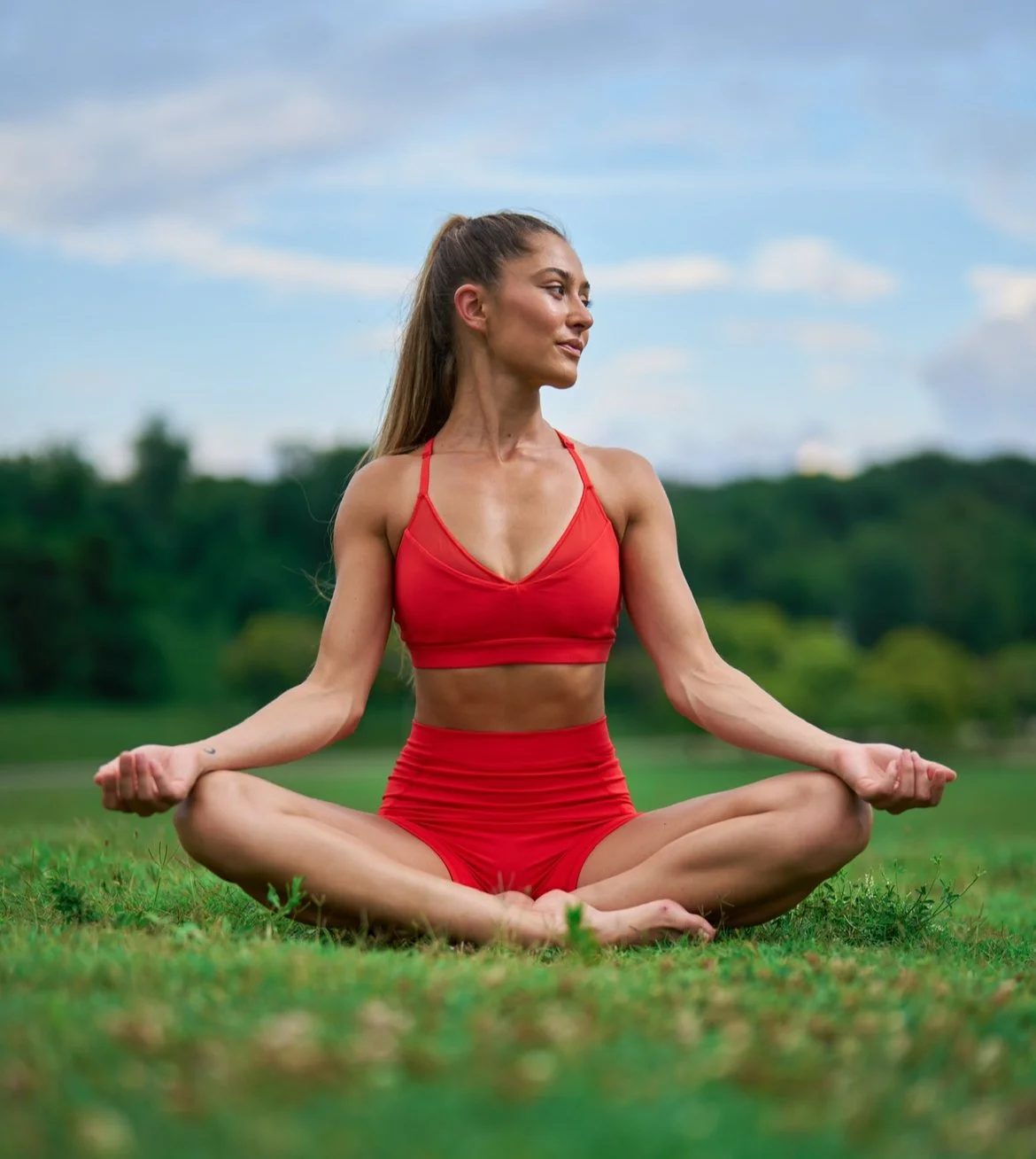 A woman practicing yoga outdoors on grass, sitting in a cross-legged meditation pose, wearing a red sports bra and matching shorts with a scenic natural background and blue sky.