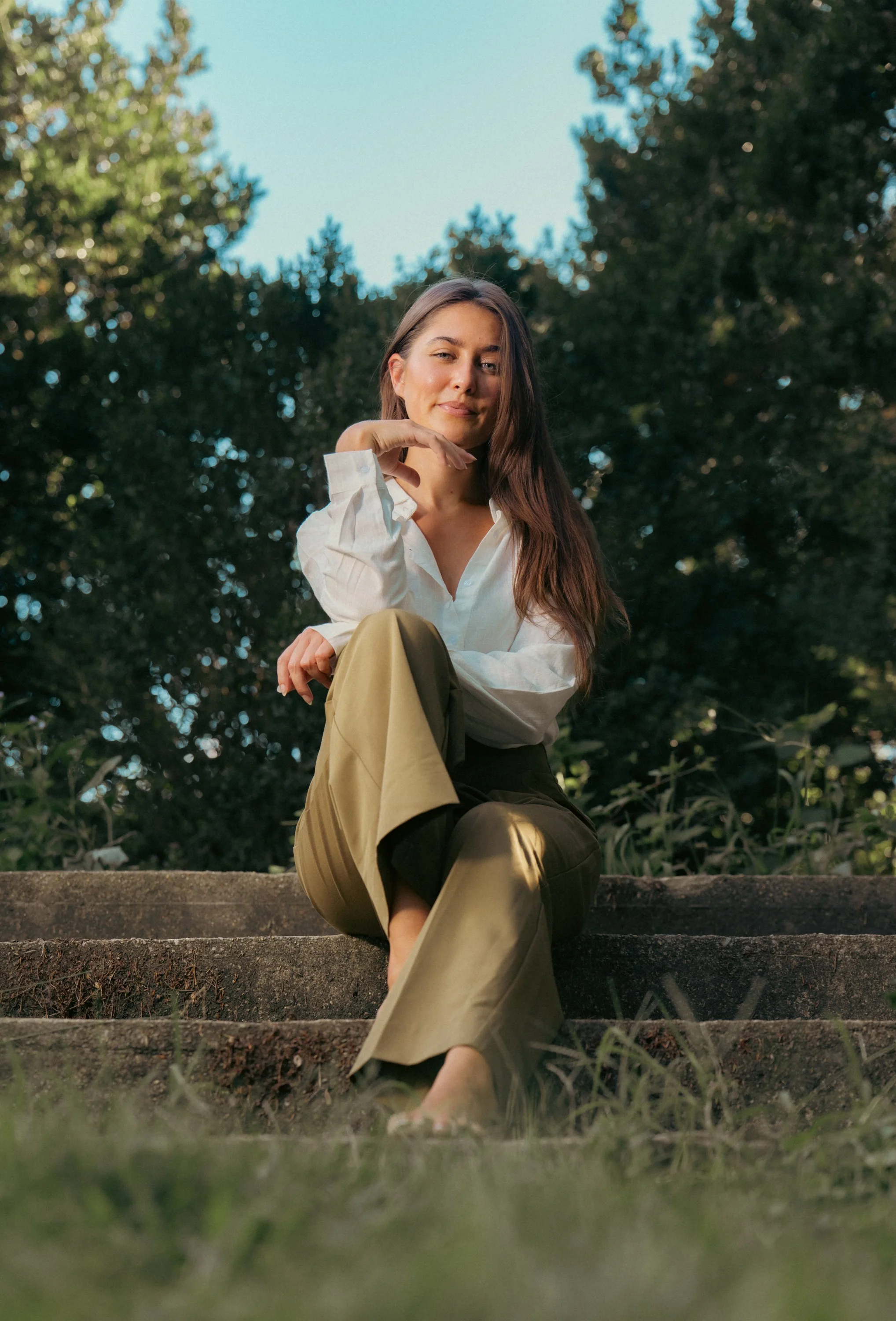 A young woman sitting on outdoor steps near trees, wearing a white blouse and beige pants, smiling softly with her hand under her chin.