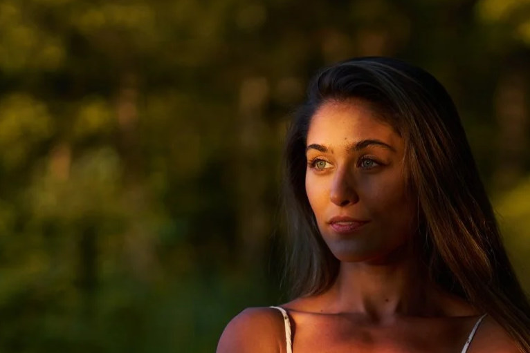 A young woman with long brown hair and blue eyes looks to the side outdoors during sunset.