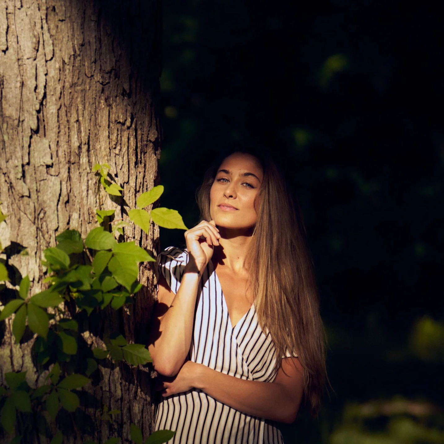A woman with long brown hair stands beside a tree in a forest, partially illuminated by sunlight, wearing a striped shirt and looking confidently at the camera.