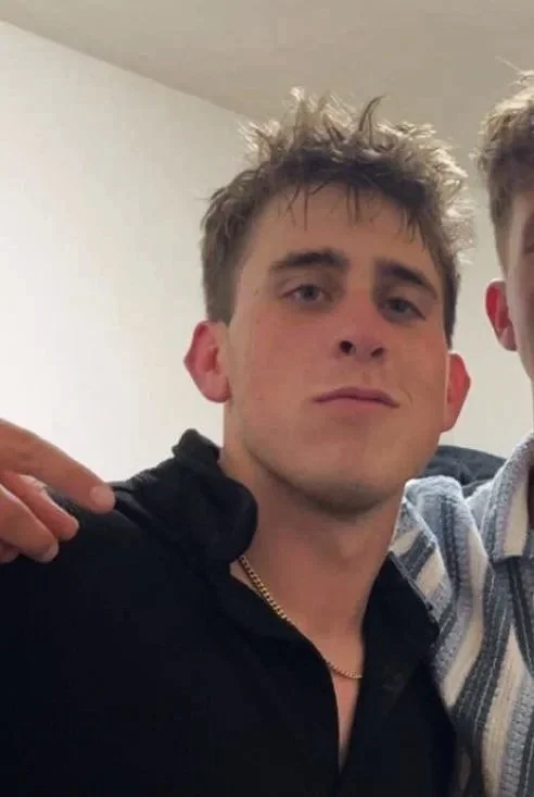 Young man with messy blond hair, wearing a black shirt and gold chain, leaning against another person partially visible on the right, indoors with plain white wall background.