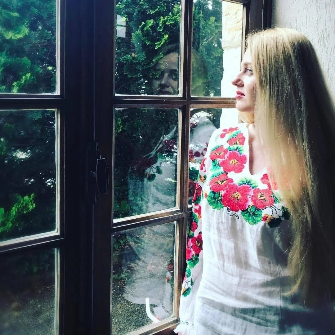 Artist Kate Sosonna standing by a window overlooking green trees.