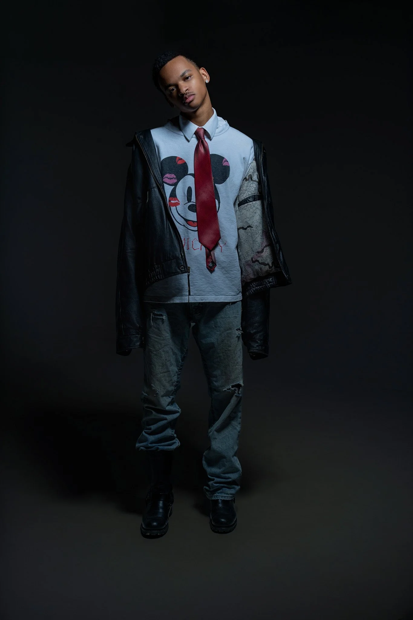 A young man standing in a dark studio, wearing a gray sweatshirt with a Mickey Mouse graphic, a red tie, a black leather jacket, distressed jeans, and black boots.