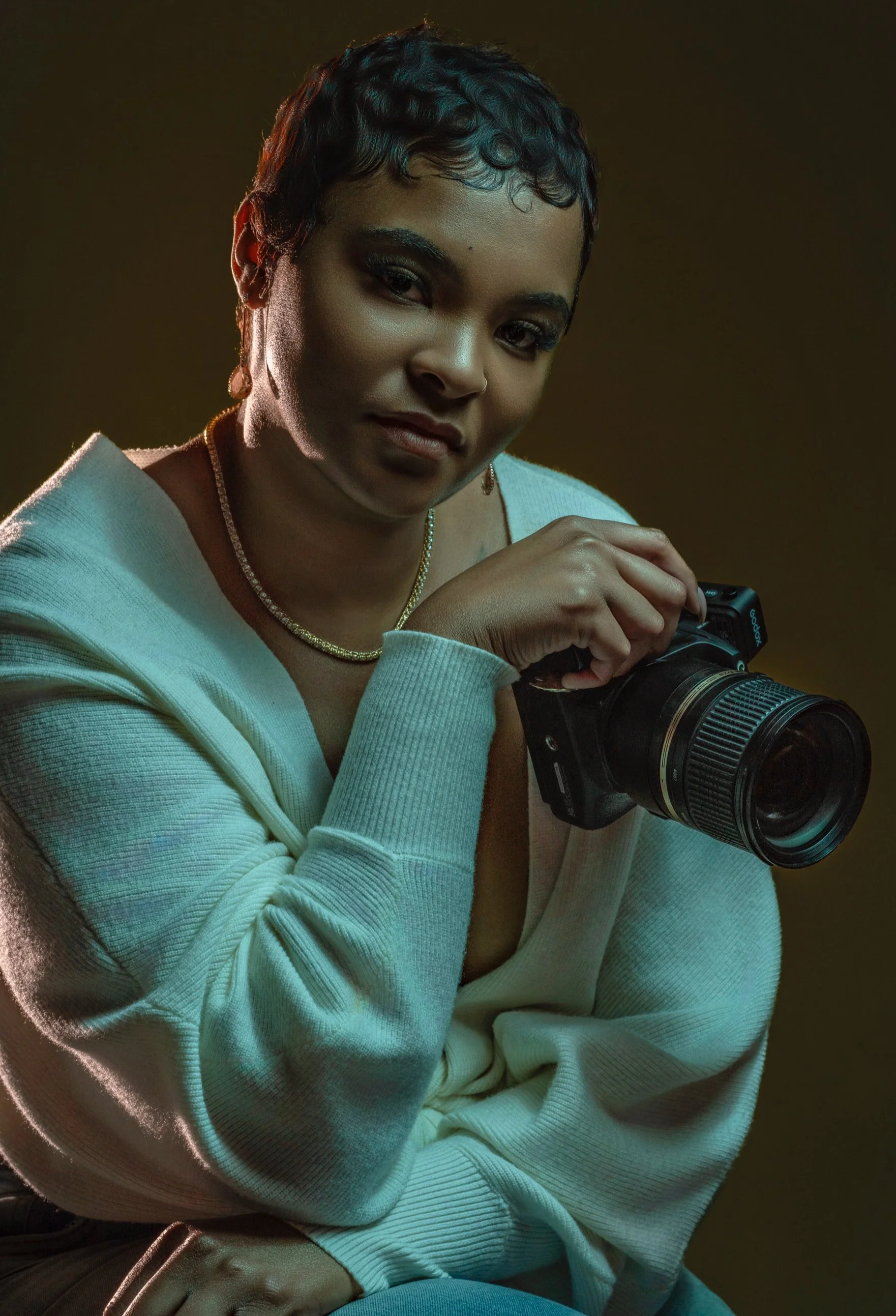 A woman with short curly hair, wearing a white hoodie and gold jewelry, holding a professional camera, looking at the camera with a slight smile in a studio with dark background.
