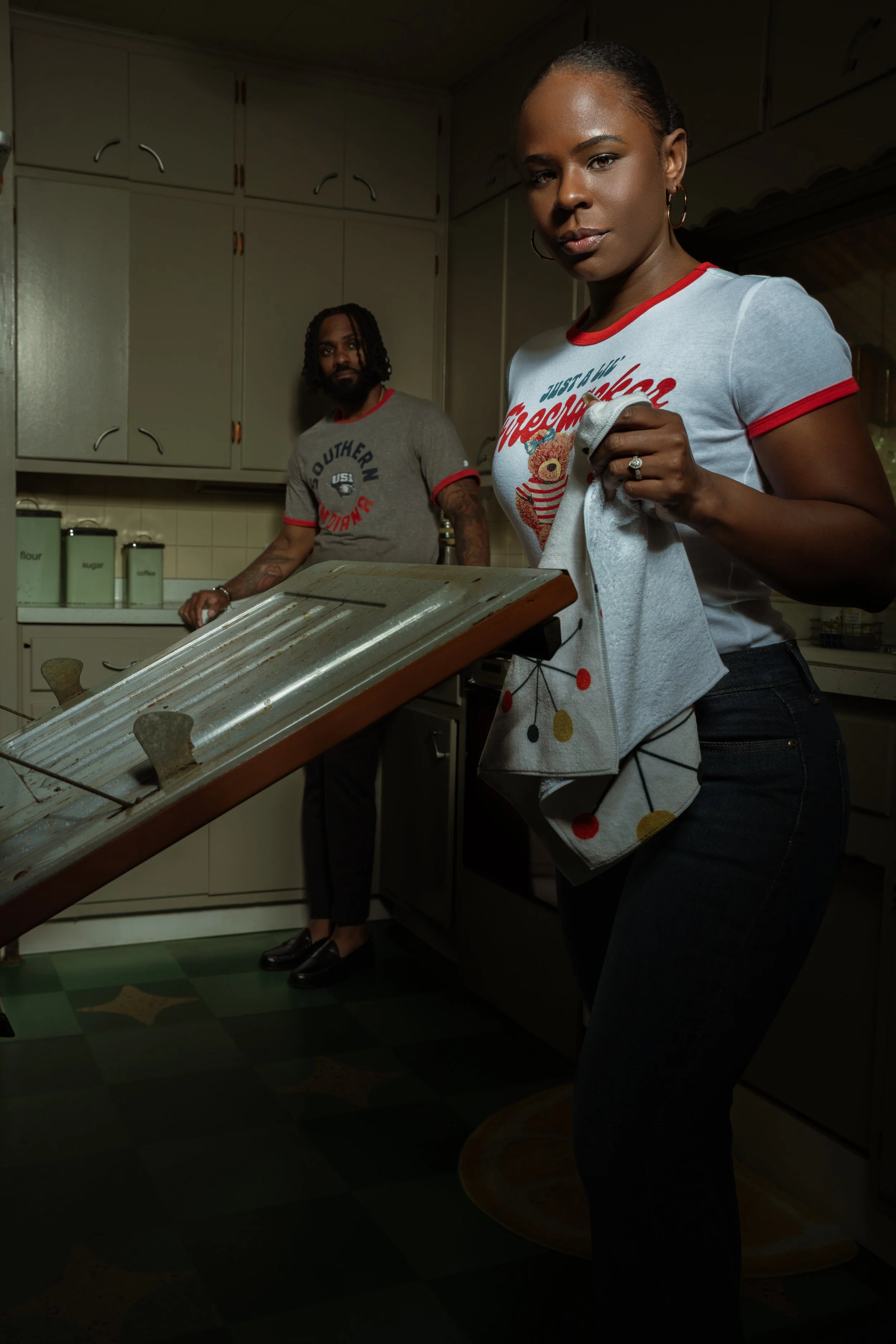 A woman with dark skin and earrings holding a kitchen towel with colorful dots, standing in a kitchen with a man with dark skin and tattoos in the background.