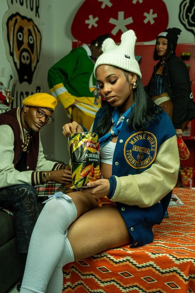 A young woman wearing a white beanie, blue and tan varsity jacket, and white thigh-high socks sitting on a patterned rug in a room with three people and colorful decor, examining a bag of Lay's potato chips.