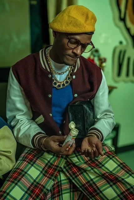 Person wearing a yellow beret, glasses, a maroon and white varsity jacket, plaid pants, and jewelry, sitting and looking at a small object in hand.