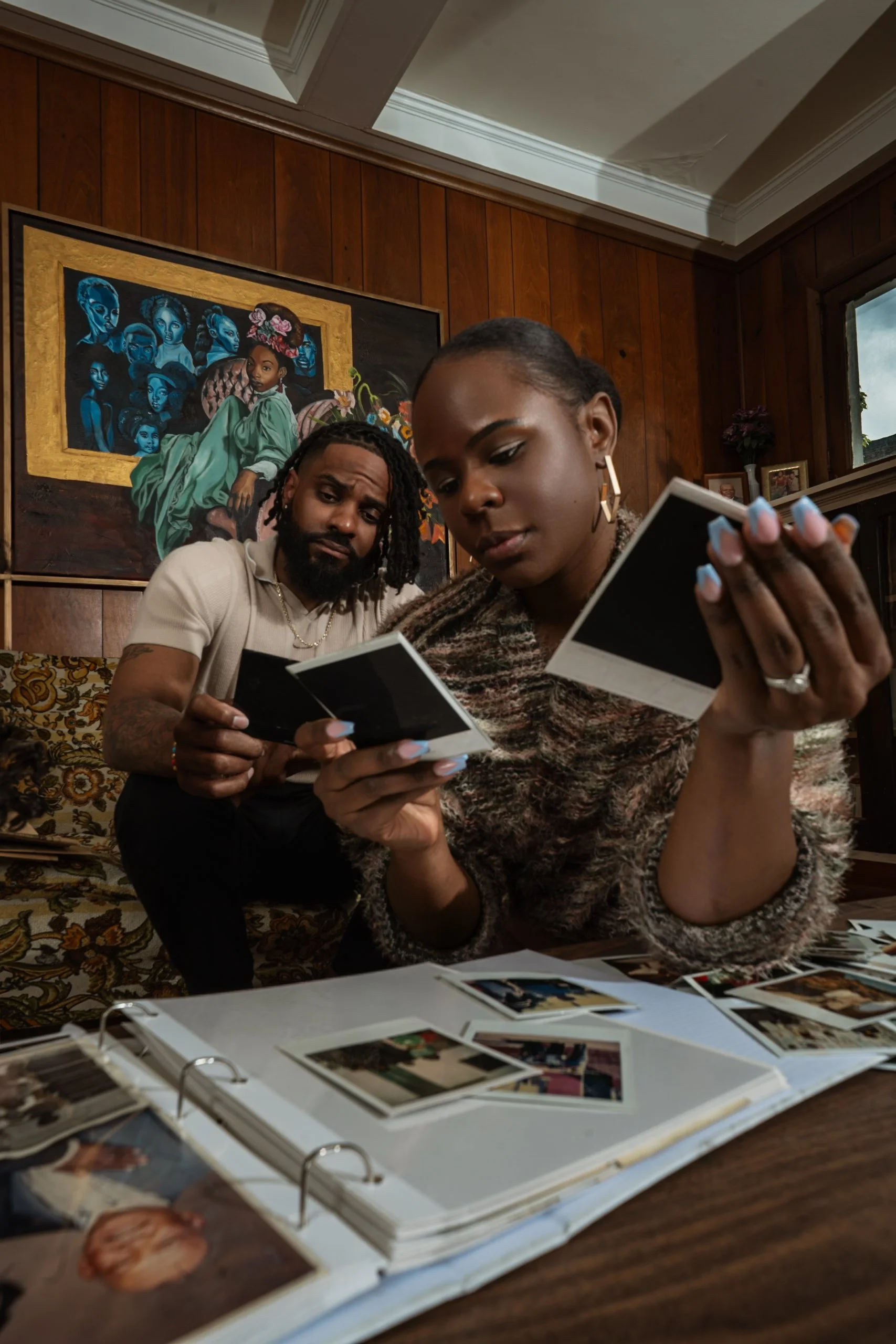 A woman and a man sitting at a table looking at photographs in an album, with a large framed painting of women on the wall behind them, and photographs on a shelf near a window.