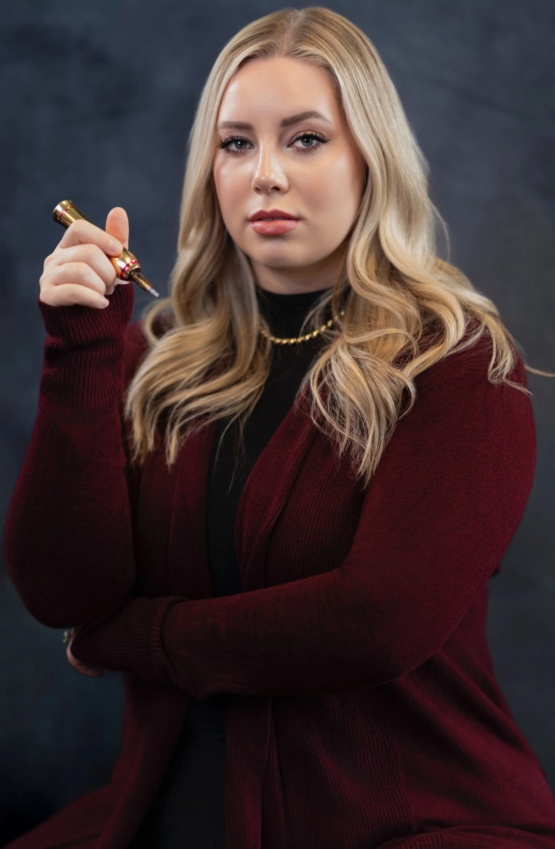 A woman with long blonde hair wearing a burgundy blazer and black turtleneck, holding a gold tattoo gun, looking at the camera against a dark gray background.