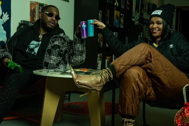 Two people sitting in a room, raising cans for a toast. The person on the left wears sunglasses and a patterned jacket, while the person on the right has a beanie, a black jacket, and brown pants. They are smiling and appear to be having a good time.