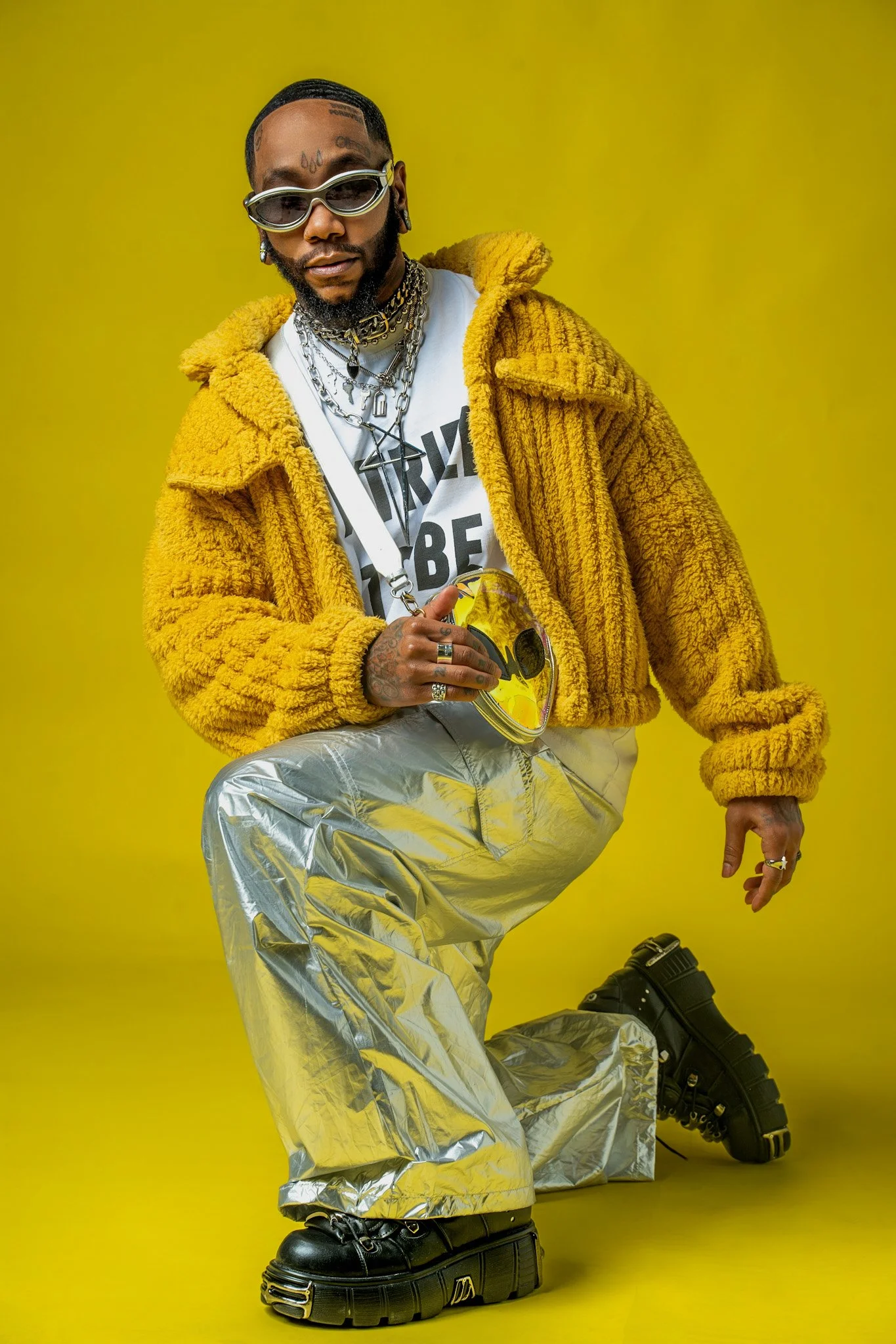 A person wearing sunglasses, a yellow furry jacket, a white T-shirt with text, shiny silver pants, and chunky black shoes, posing against a yellow background and holding a yellow mask.