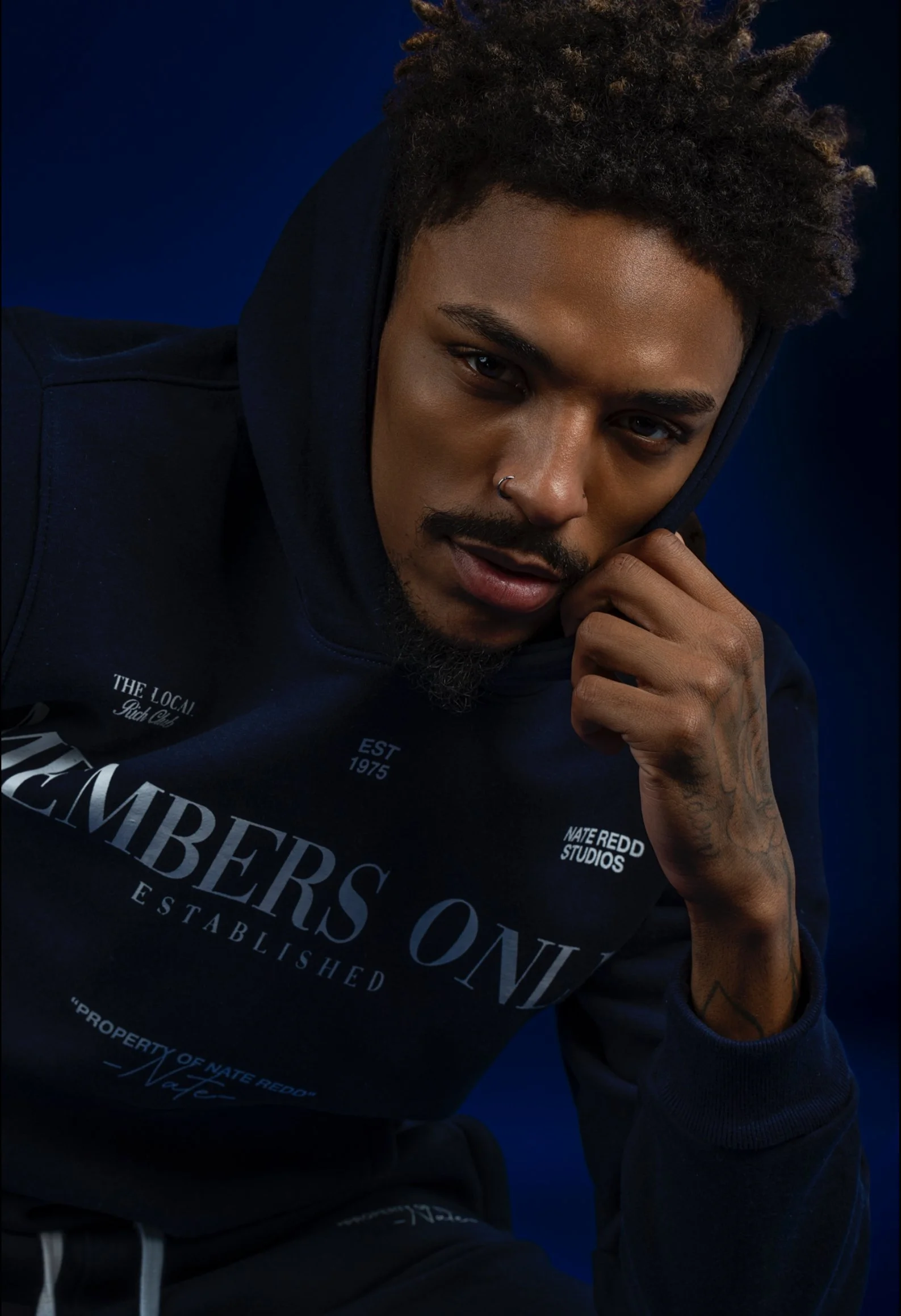A young man with curly hair and a nose ring, wearing a navy hoodie and sweatshirt, posing against a dark blue background, looking directly into the camera with a serious expression, resting his chin on his hand.
