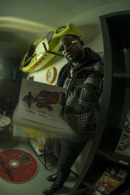 A man wearing sunglasses and a patterned jacket standing in a store or arcade with a yellow kayak hanging on the wall behind him and a mirror reflecting his image.