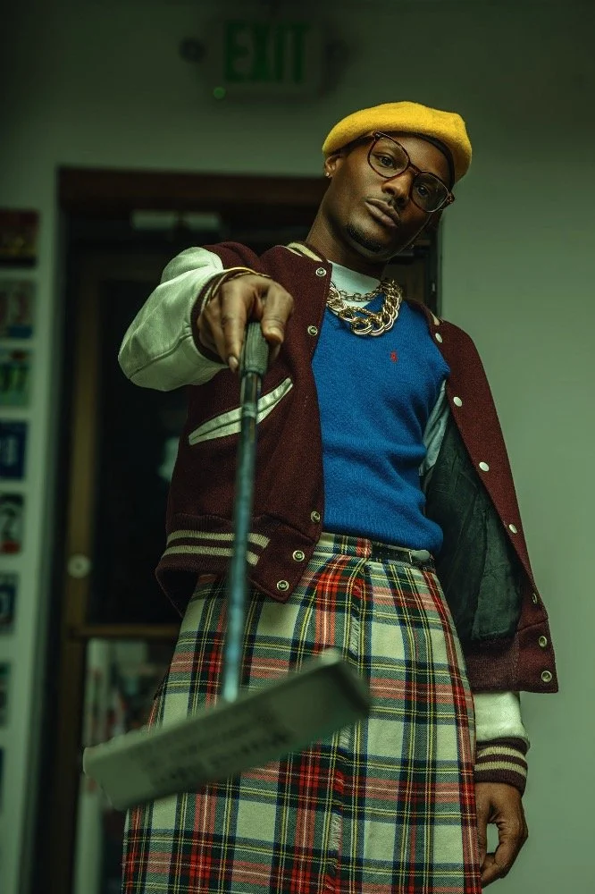 A man wearing a yellow beret, glasses, a blue shirt, a red and white letterman jacket, plaid pants, and multiple gold chains, pointing a golf club towards the camera in an indoor setting.
