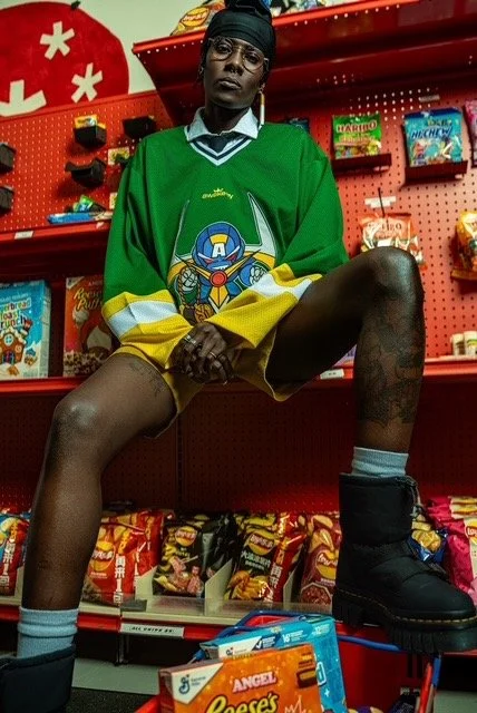 A person with short hair, glasses, and dark skin, posing in a grocery store aisle. They are wearing an oversized green hockey jersey with a large logo on the front, black boots, gray socks, and yellow shorts. The person is sitting with legs apart, one leg resting on a shopping cart filled with snacks, against a red shelf stocked with various food products and toys.