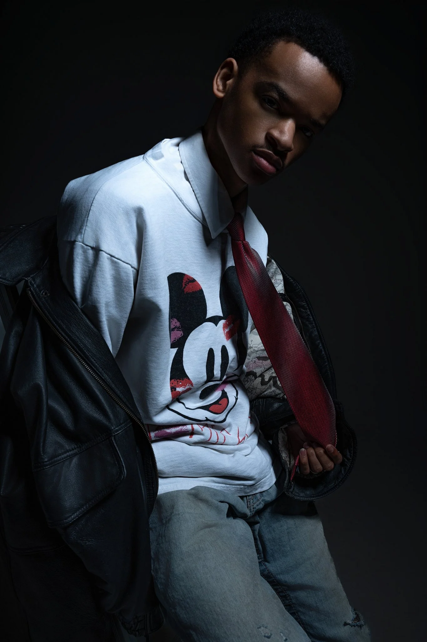 A young man with short curly hair and medium brown skin, wearing a white graphic t-shirt with a cartoon mouse face, a red tie, and a black leather jacket draped over his shoulders, poses against a dark background.