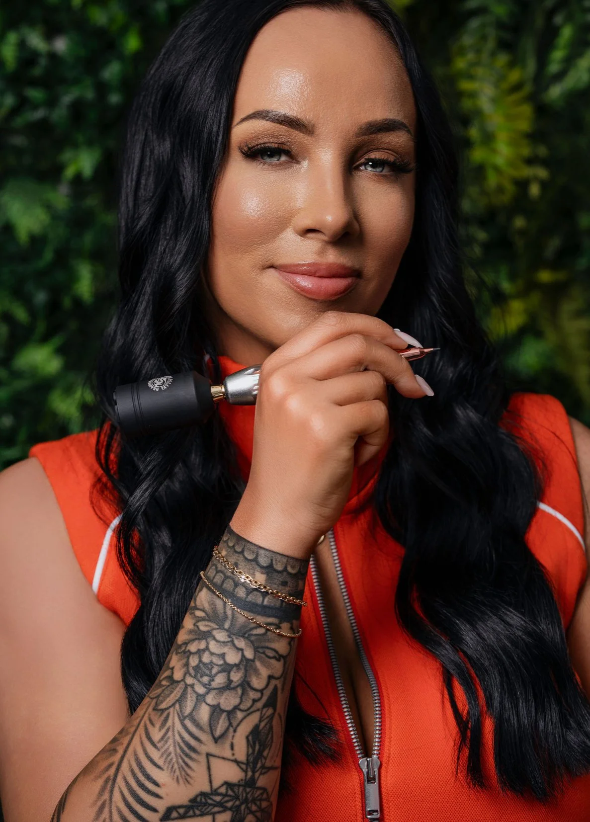 A woman with long black hair and tattoos on her arm, wearing a sleeveless orange top, holding a tattoo machine near her face, with a blurred green natural background.