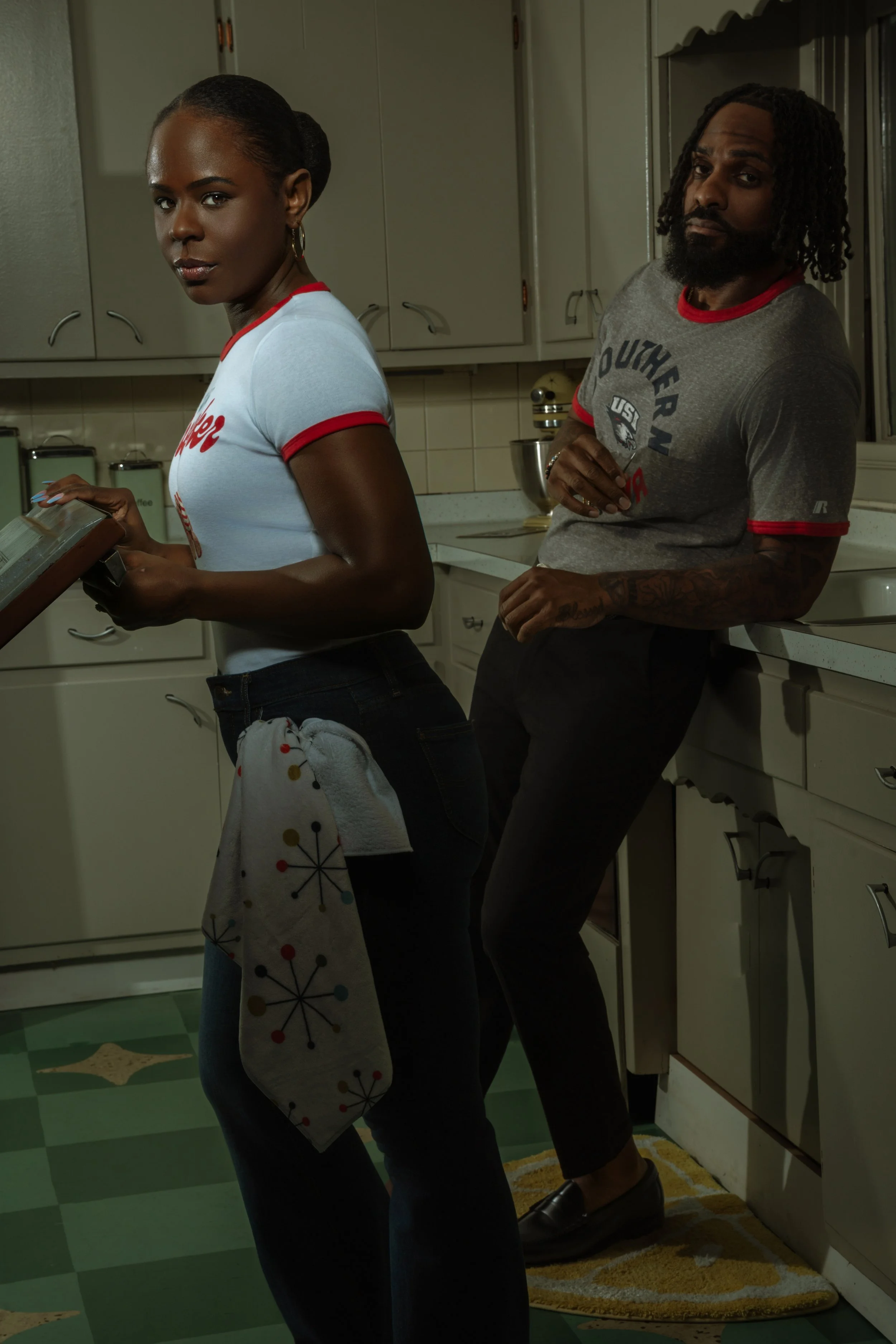 Two people standing in a kitchen, with the woman holding a book or magazine and the man leaning against the counter. The woman has dark hair styled in a bun and is wearing a white T-shirt with red trim. The man has dreadlocks, a beard, and is wearing