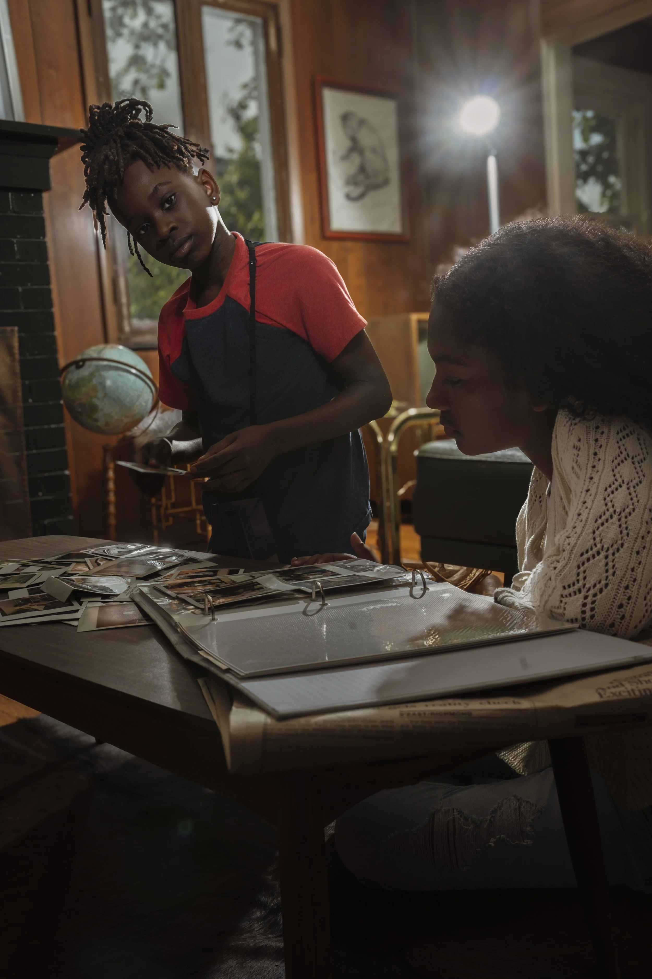 A young girl with dreadlocks looking at the camera while another girl has her head bowed, sitting at a table with photo albums and binders inside a wooden room with large windows and a bright light in the background.