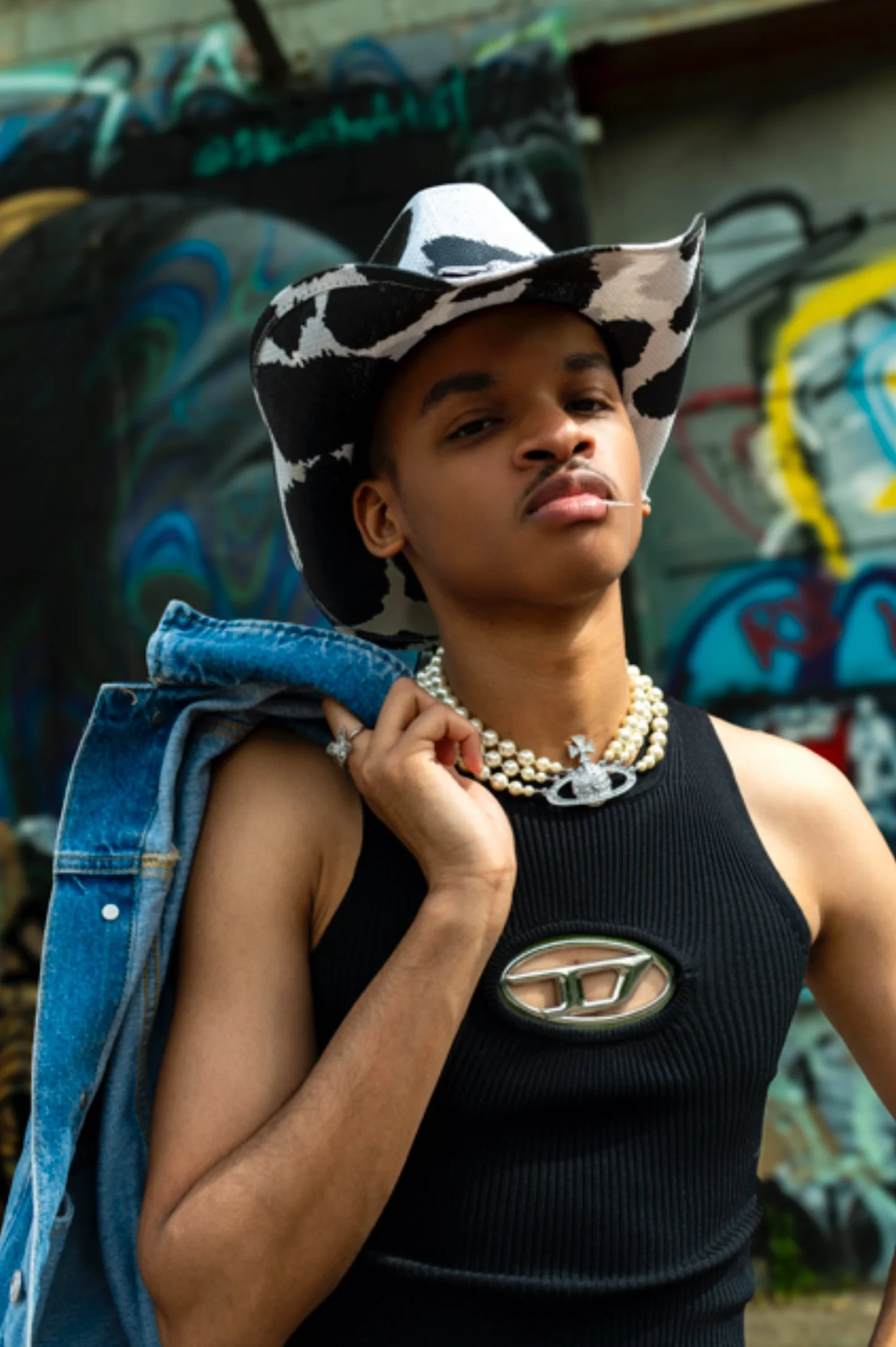A person wearing a black tank top with a logo, layered pearl necklaces, and a cow print cowboy hat, carrying a denim jacket over their shoulder, standing against a graffiti-covered wall.