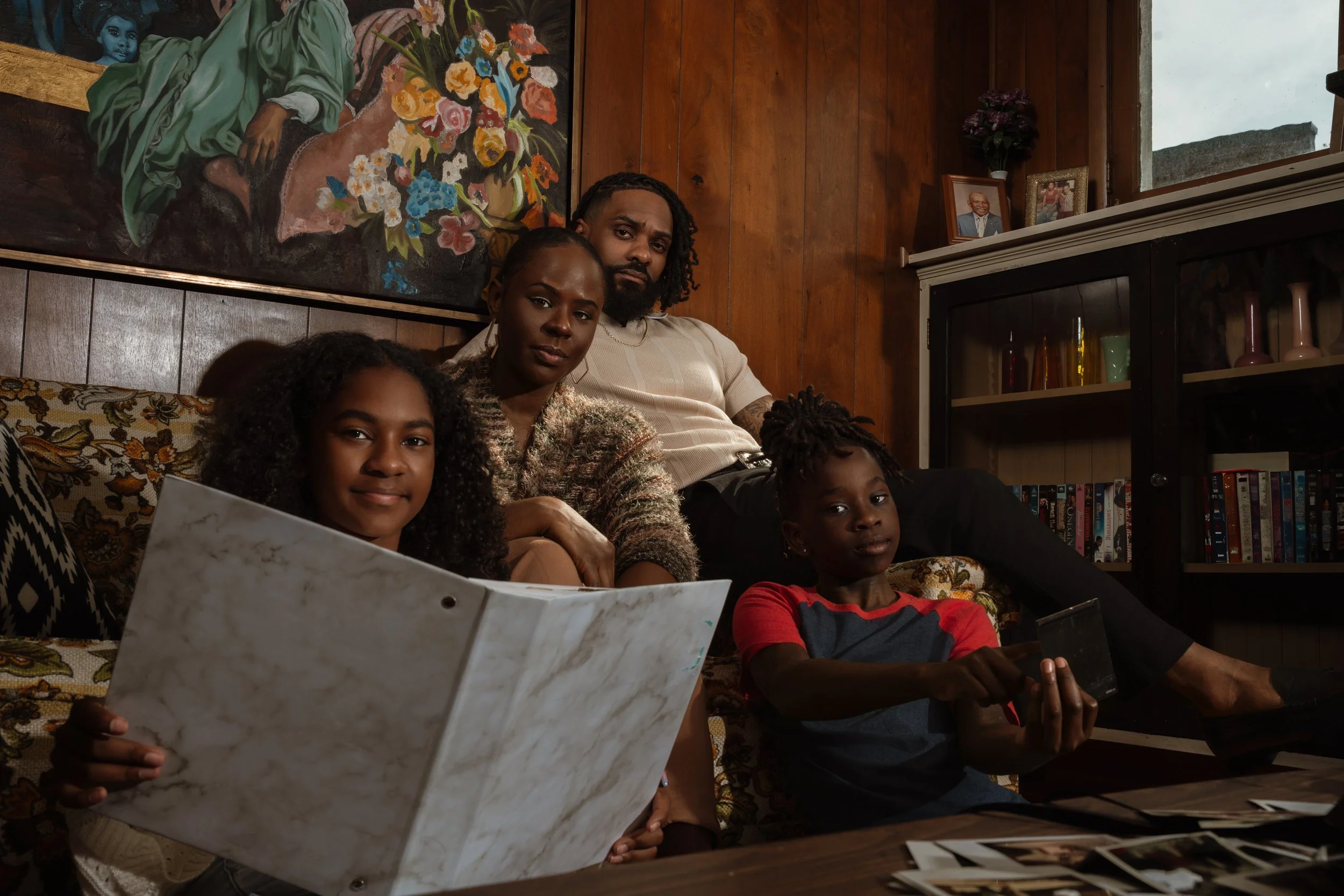 A family of four sitting on a vintage floral patterned sofa inside a wooden-paneled room. The woman and a girl are looking at a marble-patterned photo album, while the man and a boy are looking at electronic devices. The room features framed photogra