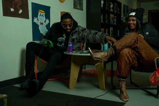 Two people seated indoors, one person has their foot resting on a table while the other is holding it. The room has colorful artwork on the walls and a bookshelf in the background. They are smiling and appear to be having a fun time.
