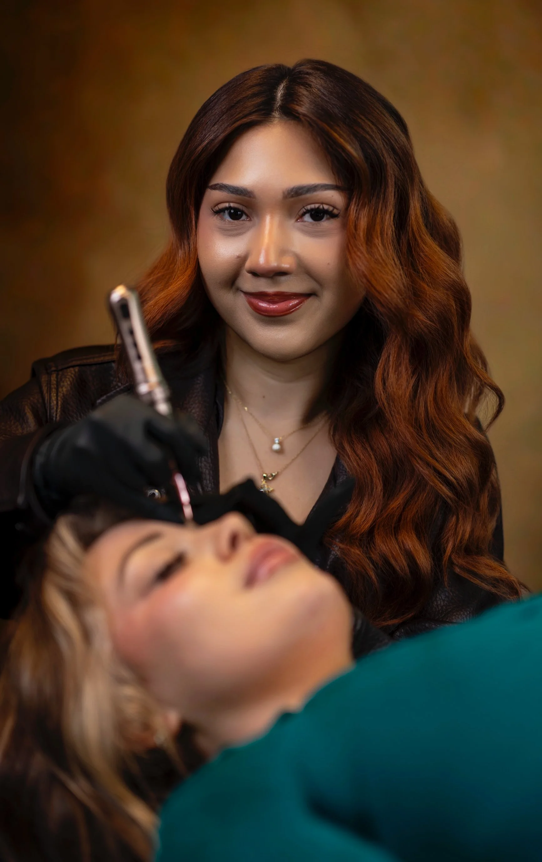 A woman with long, wavy reddish-brown hair looking at the camera, smiling, with another woman lying down in the foreground receiving a cosmetic procedure on her face, with the woman in the background holding a pen-like instrument.