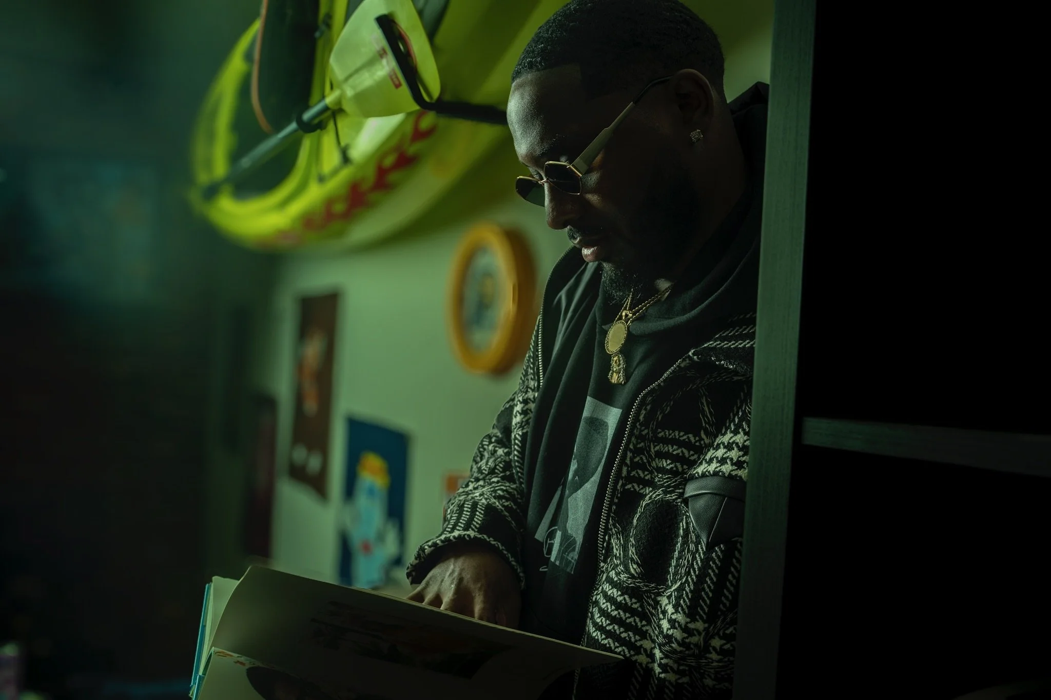A man with sunglasses and jewelry reading a book in a dimly lit room with colorful wall art.