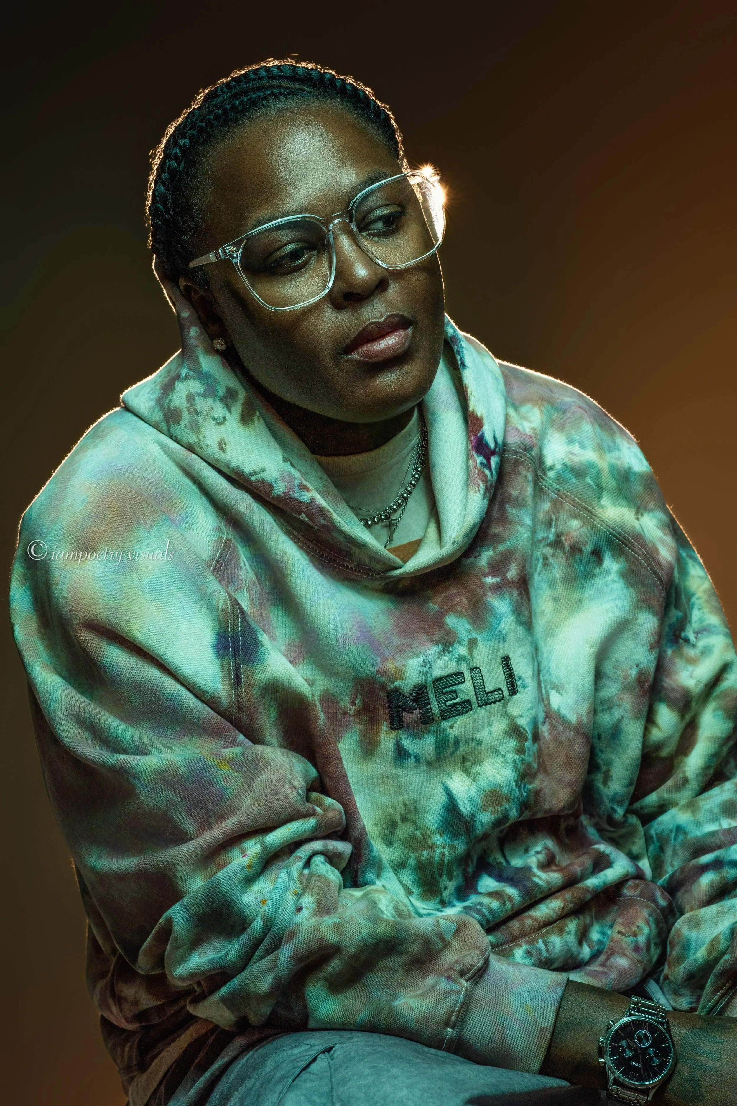 A woman with dark skin, glasses, and braided hair, dressed in a colorful tie-dye hoodie with 'MELI' written on it. She is sitting with her arms crossed, wearing a watch and a necklace, with a contemplative expression in dim lighting.