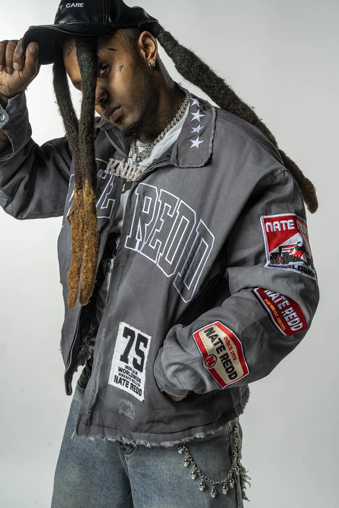 A person with long dreadlocks wearing a black cap, various patches, and a gray jacket with patches and embroidery, also wearing ripped jeans and jewelry.