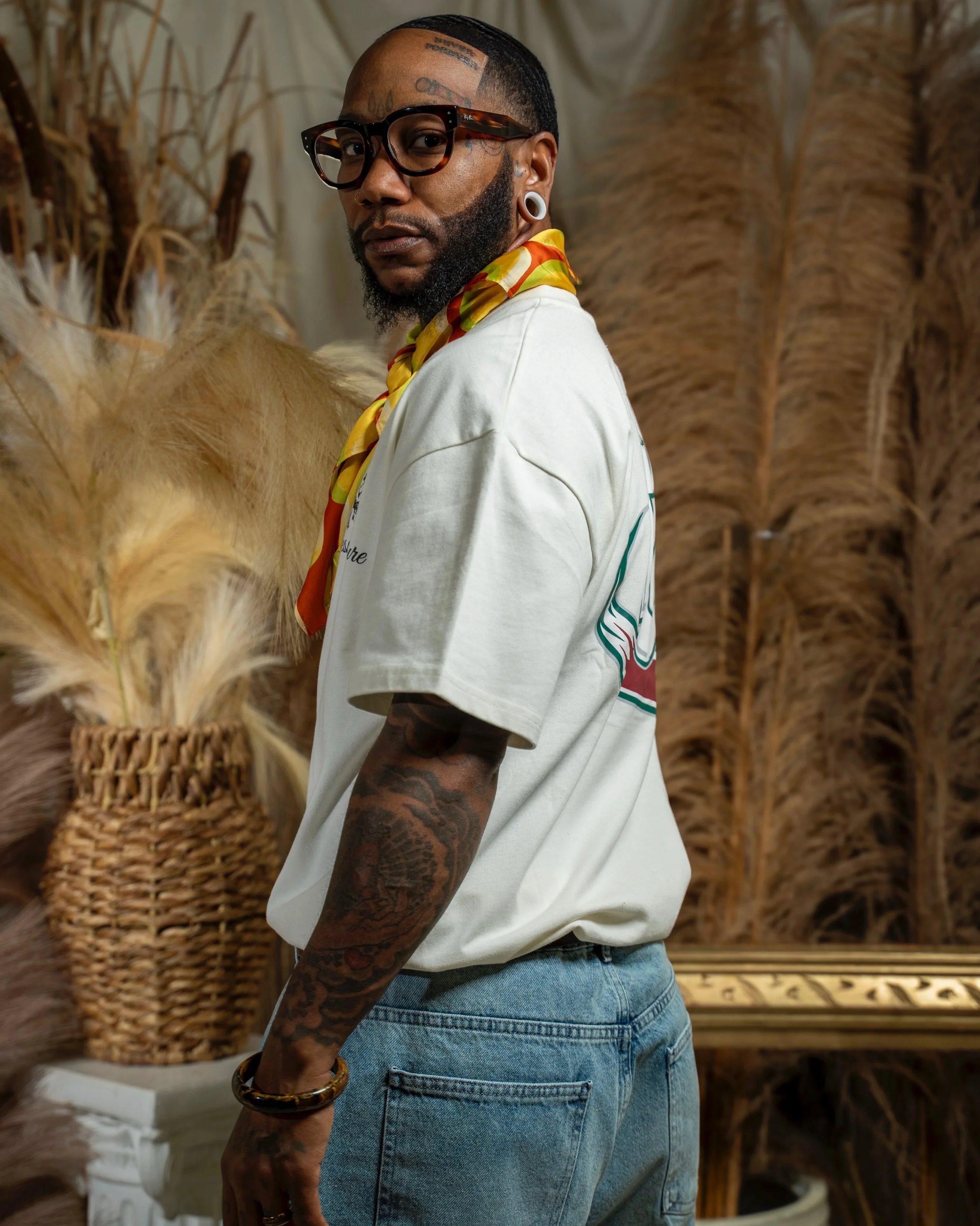 A person with tattoos, glasses, and earrings, wearing a white shirt with a colorful scarf, stands in front of a backdrop of dried plants and baskets.