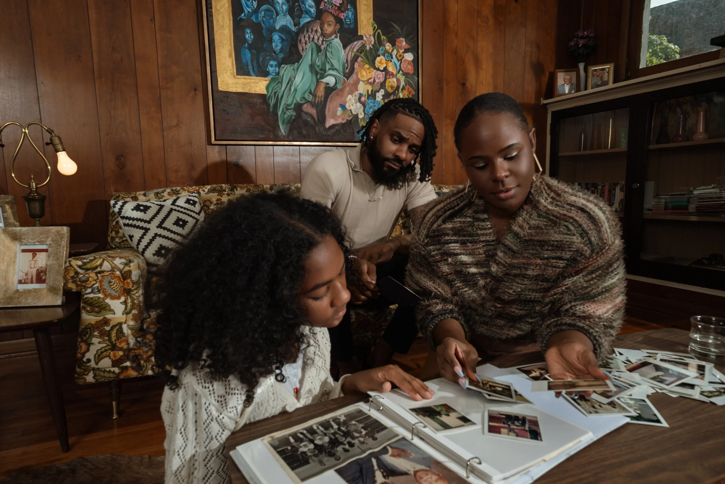 A group of four people (two children and two adults) sitting at a table looking at photo albums and pictures. The setting is a cozy room with wooden walls, decorated with artwork and family photos.