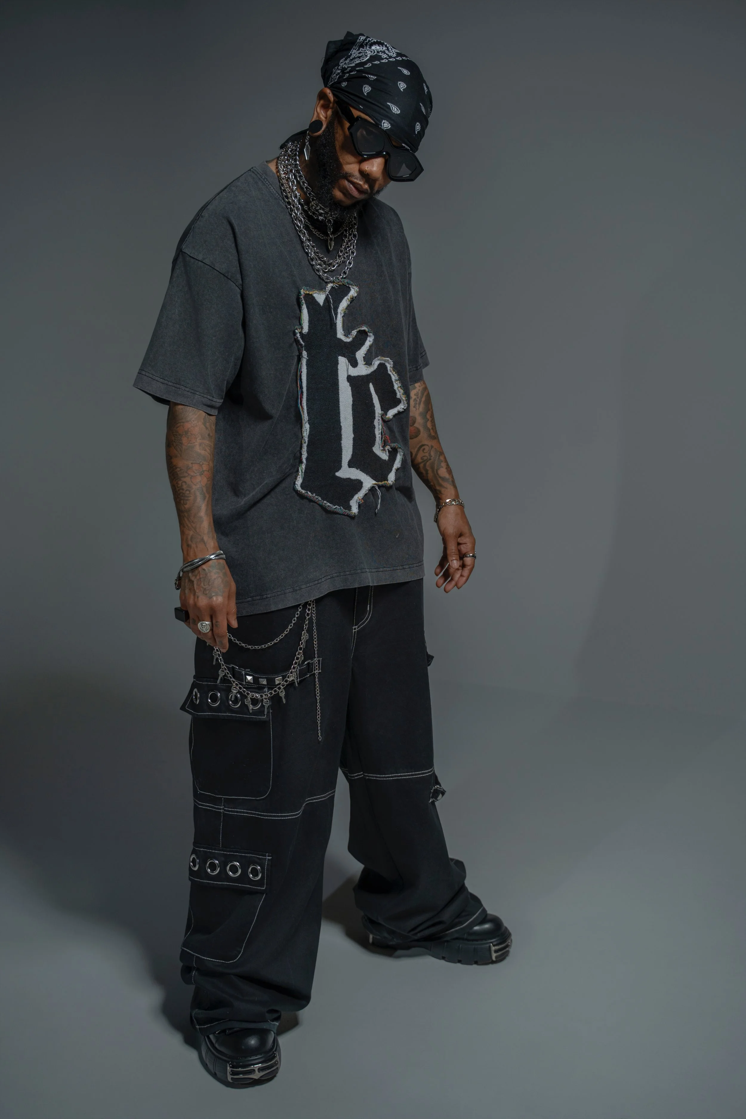 A man in black streetwear with a bandana, sunglasses, layered chains, tattoos, and chunky black shoes standing against a plain gray background.