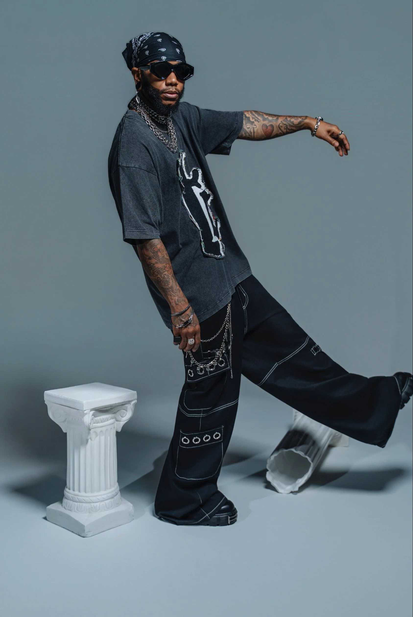 A man with sunglasses and tattoos on his arms poses next to a white, classical-style pedestal in a studio with a gray background.