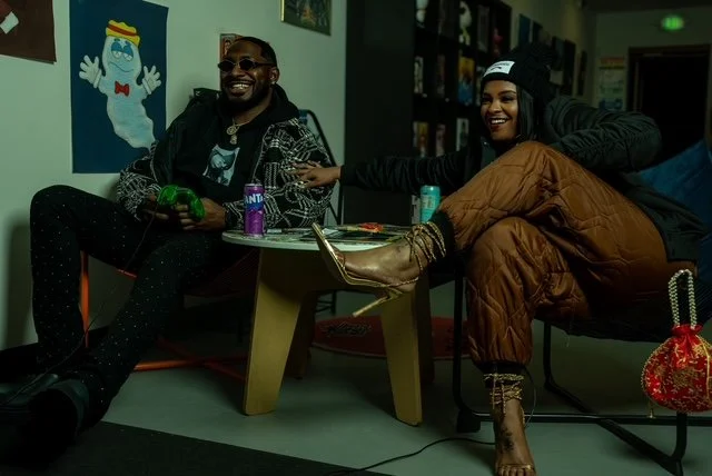 Two people sitting and laughing in a room with colorful wall art, one holding a video game controller.