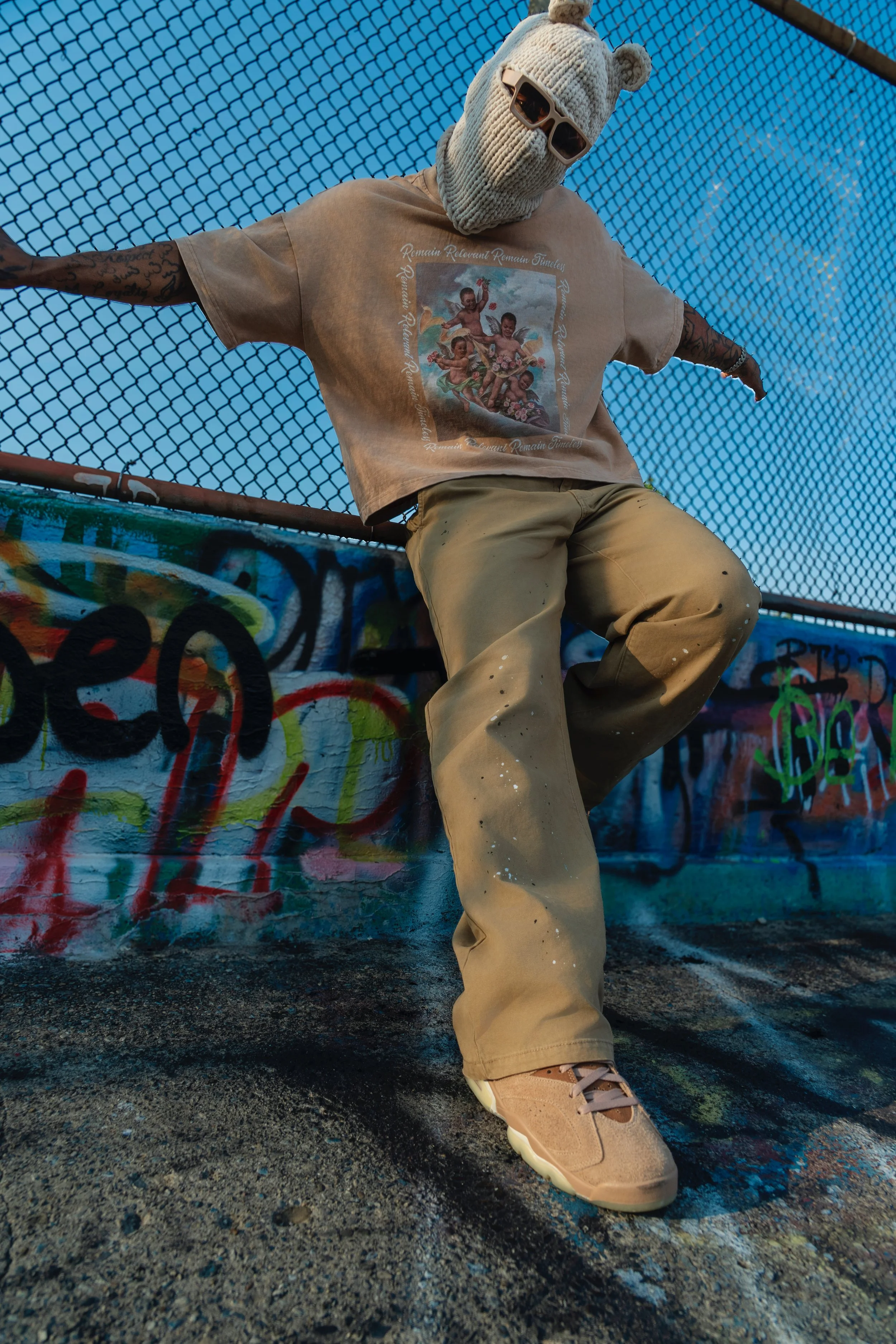 A person wearing a beige knit bear hat with ears, sunglasses, a beige T-shirt with a colorful print, beige pants with paint splatters, and beige sneakers, sitting against a chain-link fence with graffiti.