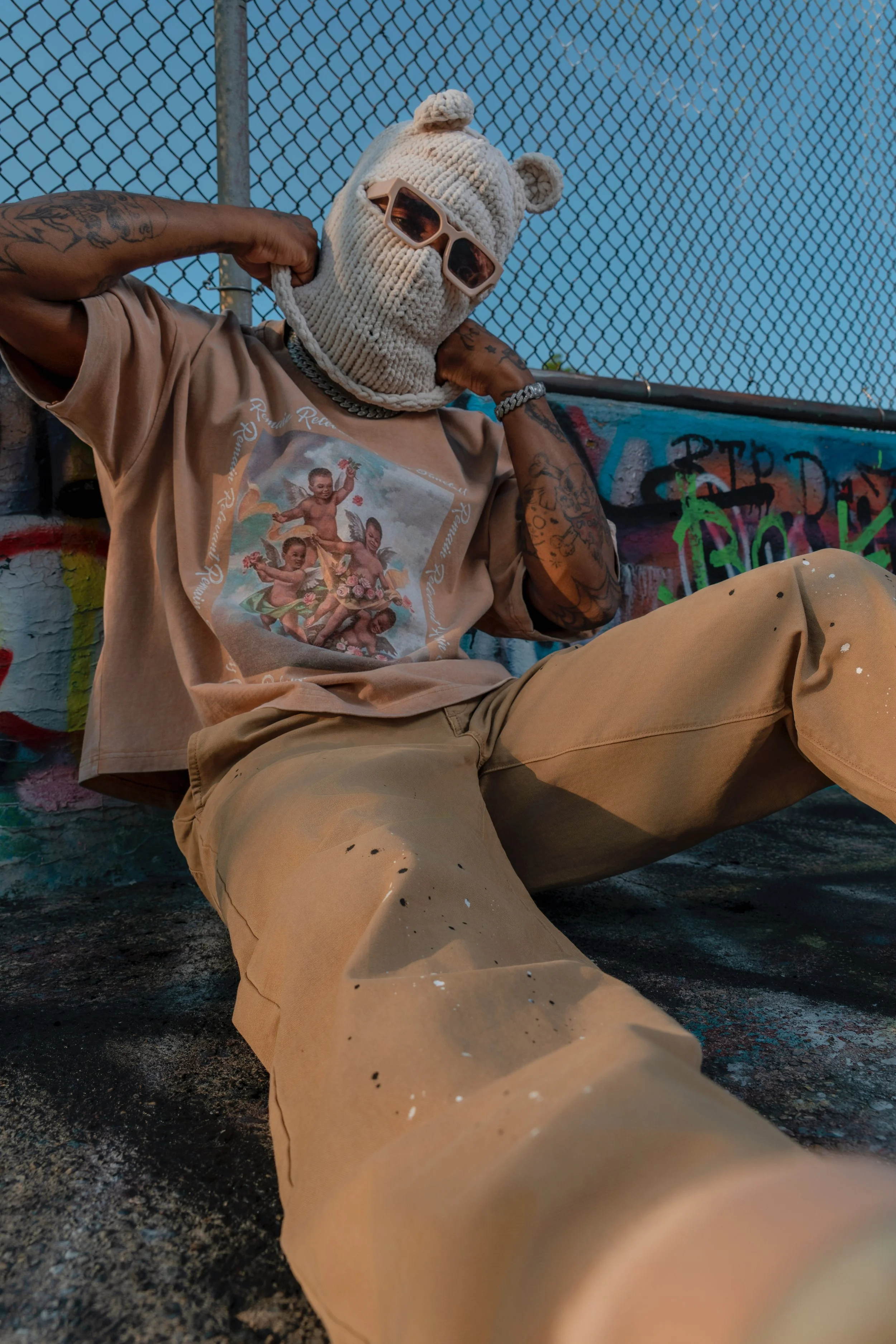 Person wearing a beige knit mask with bear ears, sunglasses, and a beige T-shirt featuring cherubs, sitting outdoors near a graffiti-covered wall and chain-link fence.
