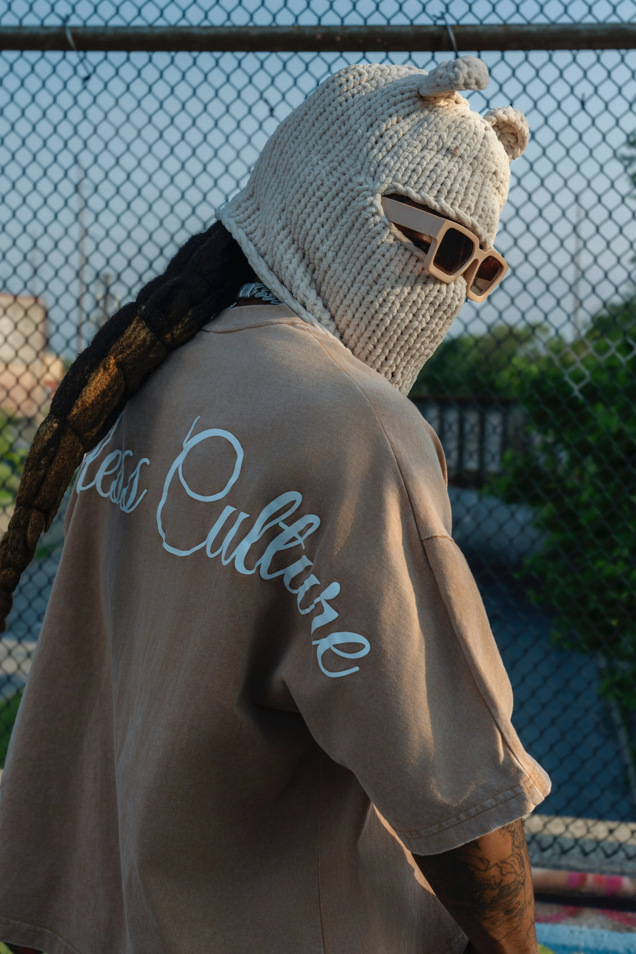 Person wearing a chunky knit hat with bear ears, sunglasses, and a beige t-shirt with white cursive text, standing outdoors near a chain-link fence during sunset.