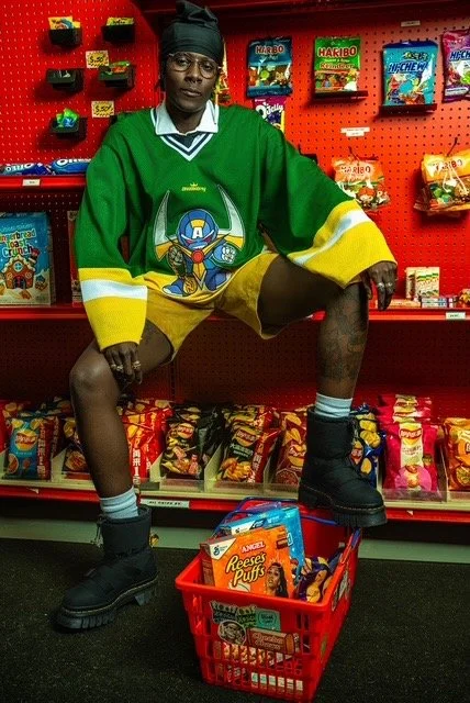 A person in a green hockey jersey and shorts, wearing black boots and a black head covering, posing inside a candy store. They are squatting with one foot on a red shopping basket filled with Reese's Puffs cereal. Behind them are shelves with various
