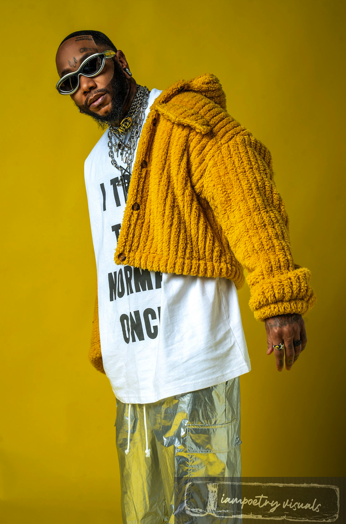 A man with dark sunglasses, multiple necklaces, and rings, wearing a yellow fuzzy jacket over a white graphic t-shirt, poses against a yellow background.