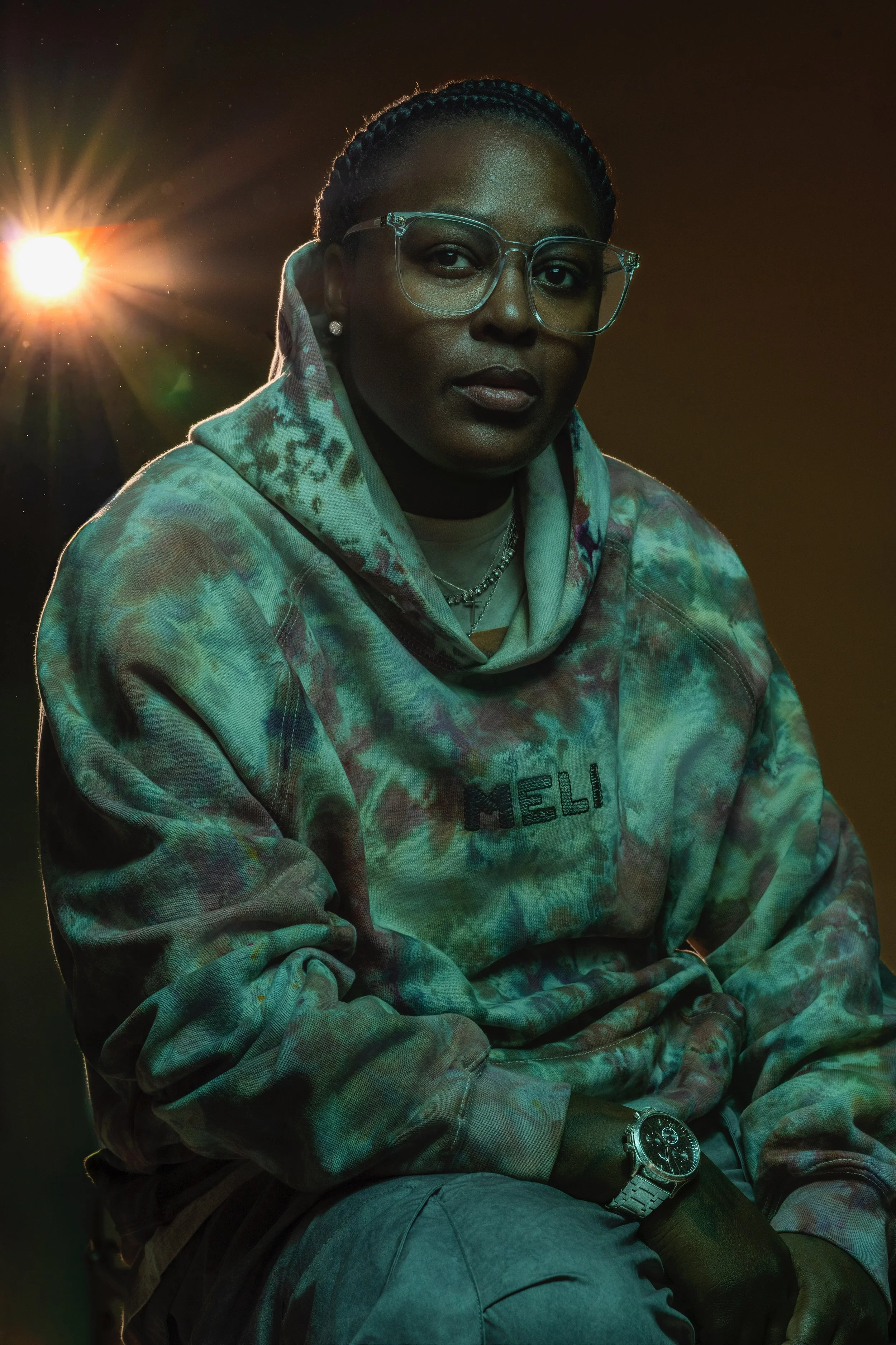 Erika wearing glasses, a tie-dye hoodie, and a watch, sitting with a serious expression, illuminated by a bright light behind her, against a dark background.